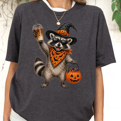 May include: A charcoal gray t-shirt featuring a graphic of a raccoon wearing a black witch hat with a small pumpkin, heart-shaped sunglasses, and an orange bandana with black bats. The raccoon holds an iced coffee and a jack-o'-lantern bucket.
