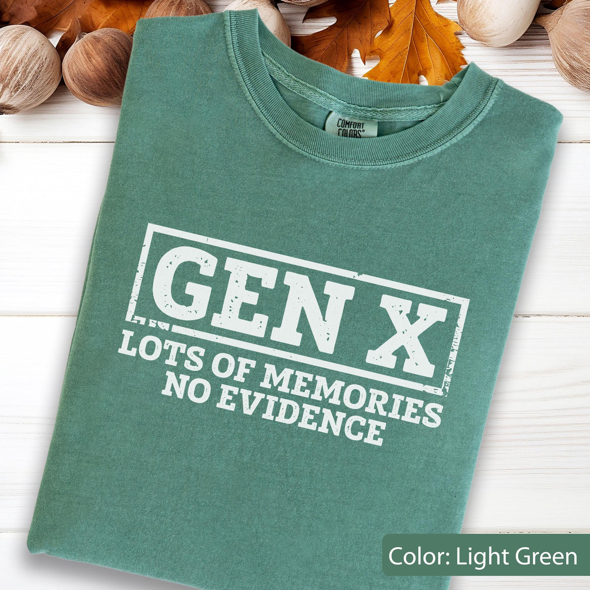 May include: A light green short-sleeve t-shirt featuring a white rectangular graphic with the text GEN X, LOTS OF MEMORIES, NO EVIDENCE. The shirt has a crew neckline and a Comfort Colors brand label visible on the inside of the collar area.