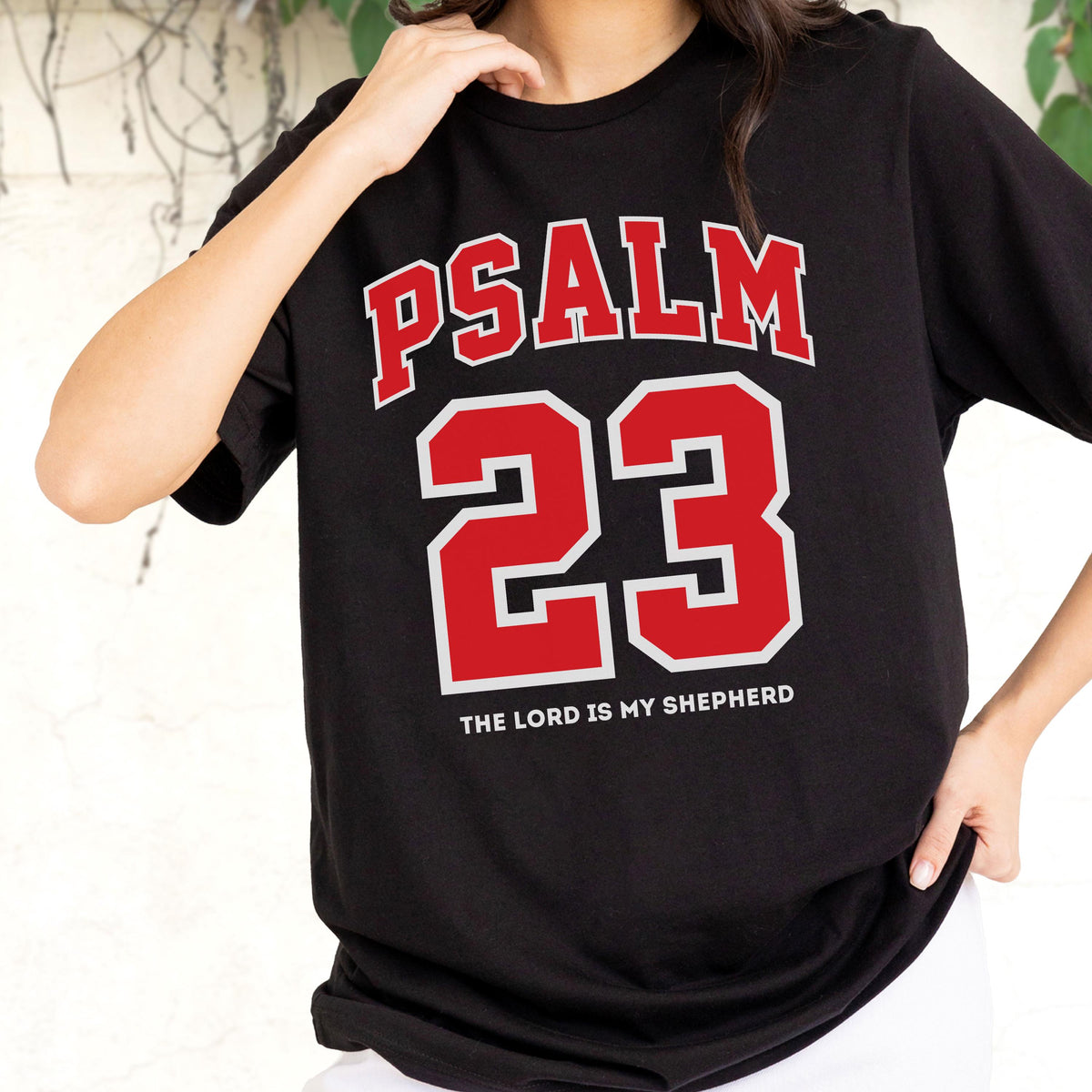 May include: A black short-sleeved t-shirt featuring the red text PSALM 23 in an arched varsity font above a large red number 23 with a white outline. Below the number, the text THE LORD IS MY SHEPHERD is printed in a smaller white font.
