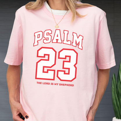 May include: A light pink short-sleeved t-shirt featuring the red text PSALM 23 in a collegiate font, with the number 23 in large red outlined block numerals and the text THE LORD IS MY SHEPHERD printed in smaller red letters underneath.