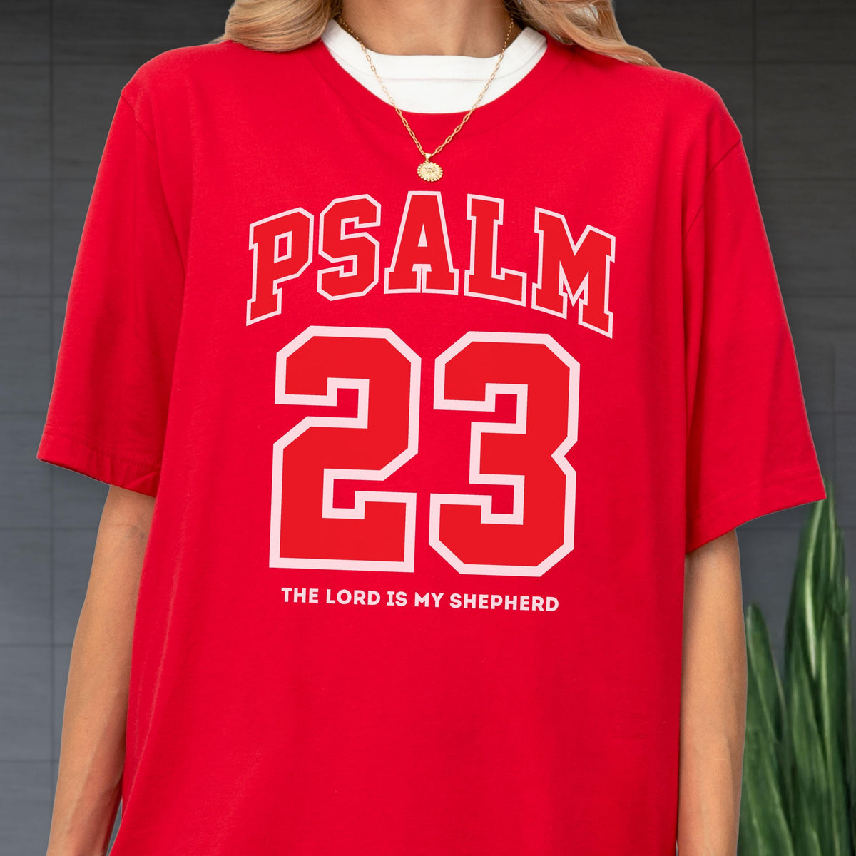 May include: A bright red short-sleeved t-shirt featuring the white text PSALM in an arched font above the large white number 23. Below the number, the text THE LORD IS MY SHEPHERD is printed in white. A gold chain necklace is worn over the shirt.