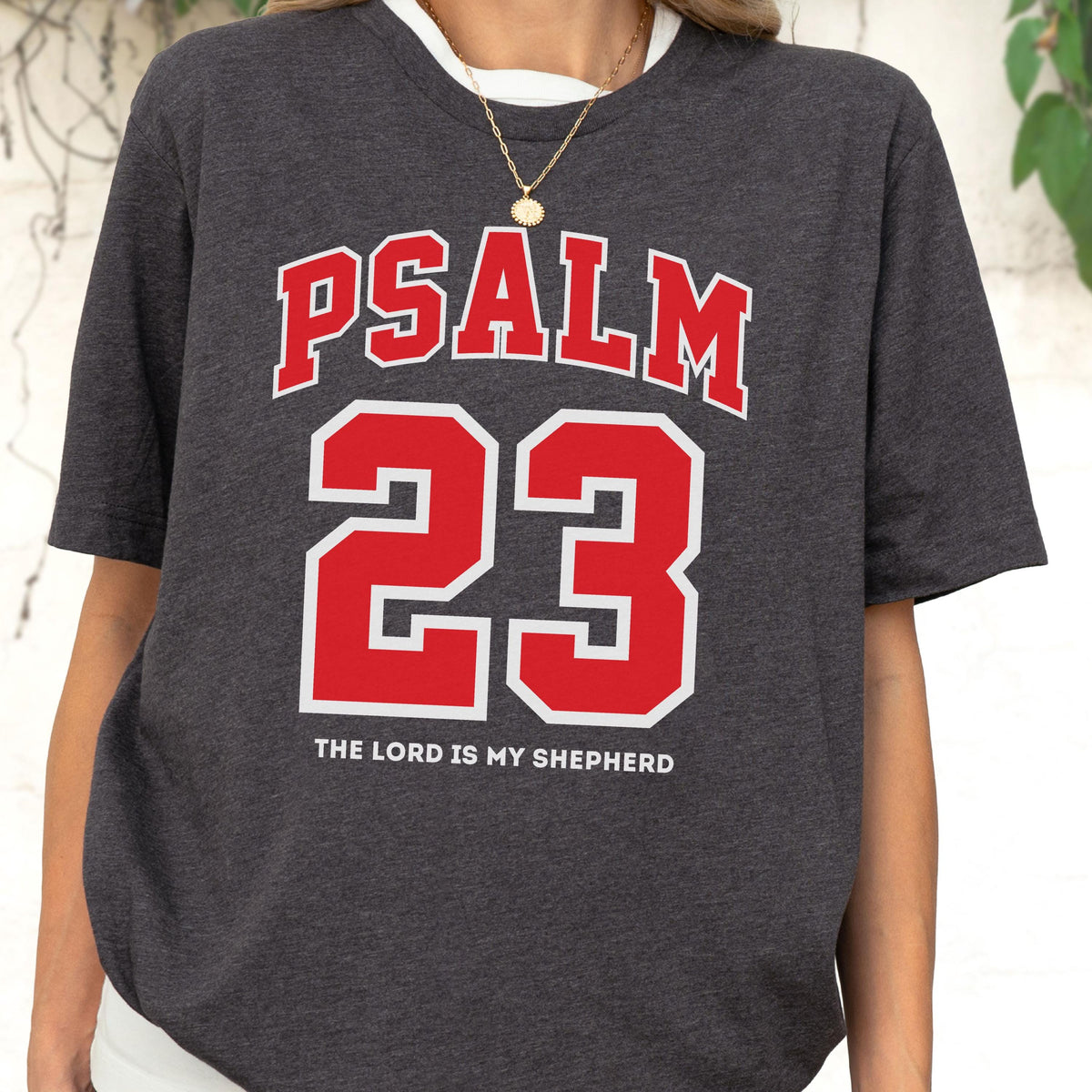 May include: A dark charcoal gray short-sleeve t-shirt featuring the red text PSALM in an arched font above the large red number 23. Below the number, the white text reads THE LORD IS MY SHEPHERD. The shirt has a crew neckline and a heathered texture.