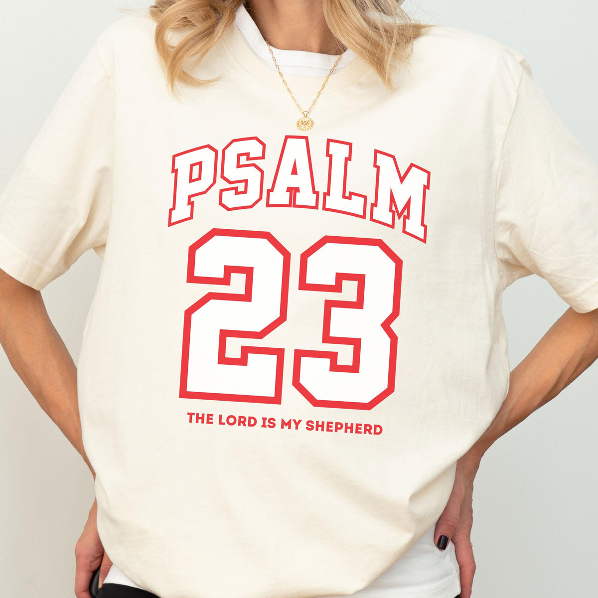 May include: A cream-colored short-sleeve t-shirt featuring red text that reads PSALM 23, a large red outlined number 23, and the text THE LORD IS MY SHEPHERD in red. The shirt has a crew neckline and a relaxed fit, worn with a gold chain necklace.