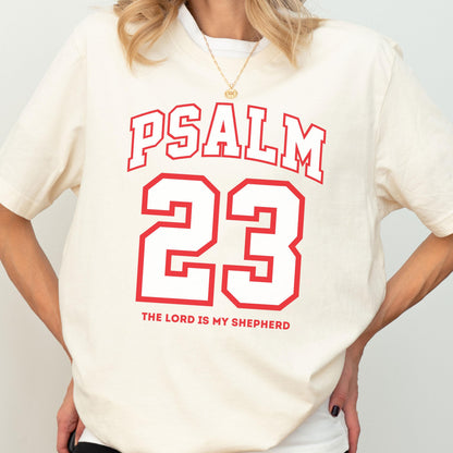 May include: A cream-colored short-sleeve t-shirt featuring red text that reads PSALM 23, a large red outlined number 23, and the text THE LORD IS MY SHEPHERD in red. The shirt has a crew neckline and a relaxed fit, worn with a gold chain necklace.