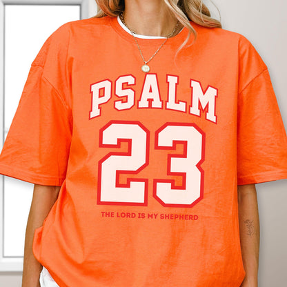 May include: An orange short-sleeved t-shirt featuring the white text PSALM 23 in a collegiate font above a large white number 23 with a red outline. Below the number, the text THE LORD IS MY SHEPHERD is printed in a smaller red sans-serif font.