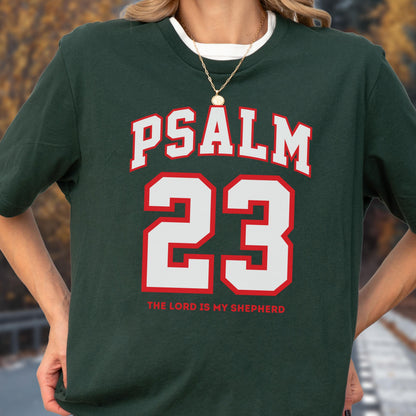 May include: A dark green short-sleeve t-shirt featuring the white text PSALM in an arched font above the large red and white number 23. Below the number, the text THE LORD IS MY SHEPHERD is printed in red capital letters on the front of the shirt.