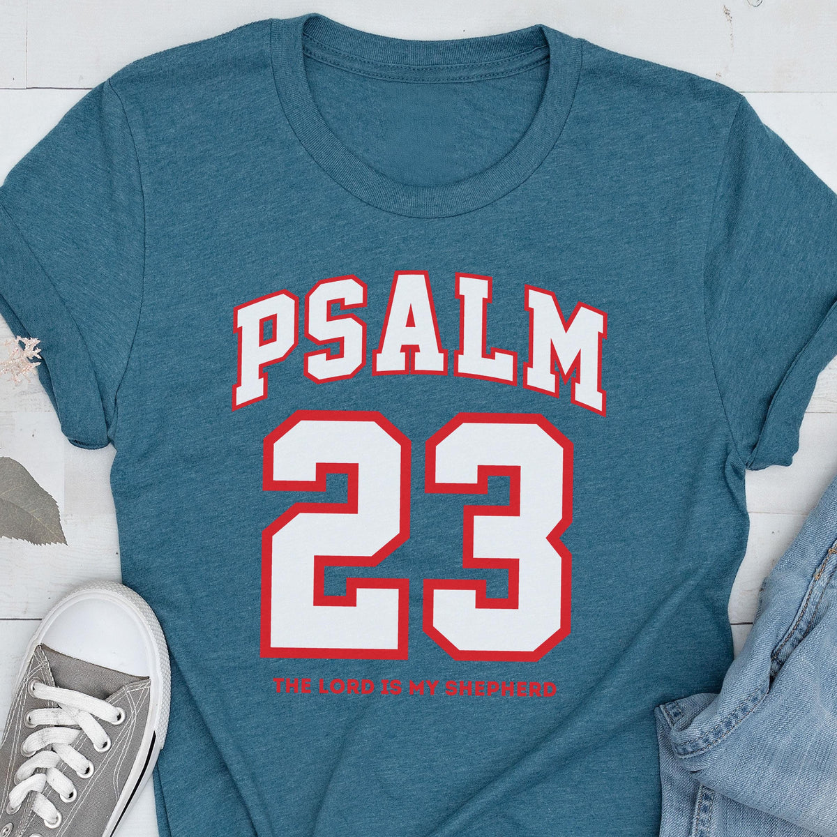 May include: A heathered teal blue short-sleeve t-shirt featuring the white text PSALM 23 in a collegiate font above the large white number 23 with a red outline. Below the number, the text THE LORD IS MY SHEPHERD is printed in small red letters.