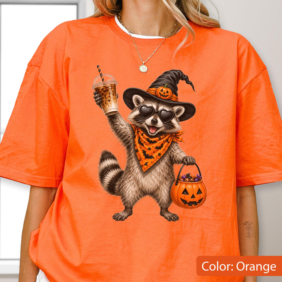 May include: An orange t-shirt featuring a graphic of a raccoon wearing a black witch hat with a small pumpkin, heart-shaped sunglasses, and an orange bandana with a black bat pattern. The raccoon holds a plastic iced coffee and a pumpkin bucket.