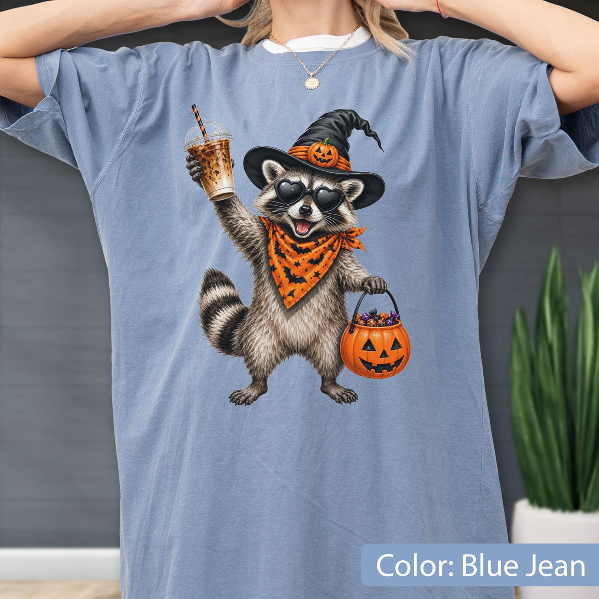May include: A blue jean colored t-shirt featuring a graphic of a raccoon wearing a witch hat with a pumpkin, heart-shaped sunglasses, and an orange bandana with bats. The raccoon holds a plastic cup and a jack-o'-lantern bucket filled with candy.