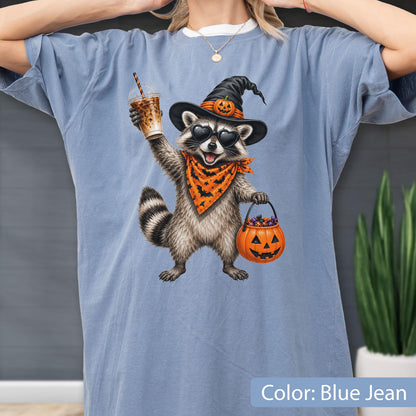 May include: A blue jean colored t-shirt featuring a graphic of a raccoon wearing a witch hat with a pumpkin, heart-shaped sunglasses, and an orange bandana with bats. The raccoon holds a plastic cup and a jack-o'-lantern bucket filled with candy.