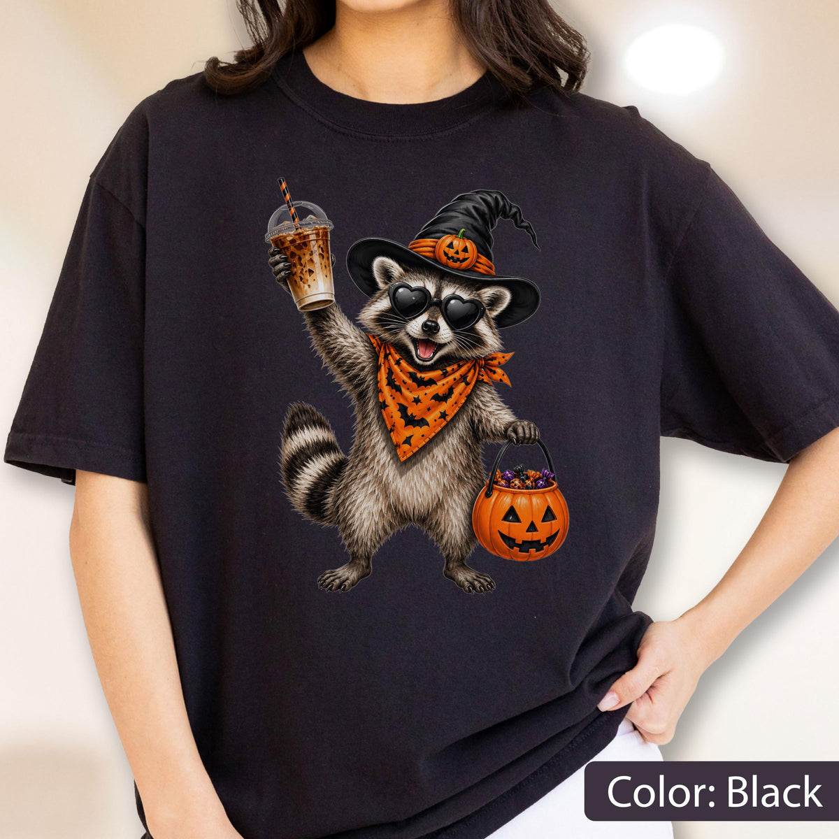 May include: A black t-shirt featuring a graphic of a raccoon wearing a witch hat with a small pumpkin, heart-shaped sunglasses, and an orange bandana with a black bat pattern. The raccoon holds an iced coffee and a jack-o'-lantern candy bucket.