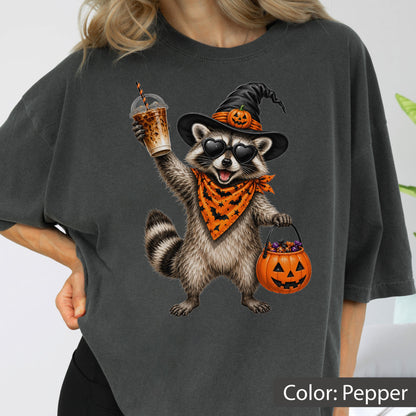 May include: A charcoal gray t-shirt featuring a graphic of a raccoon wearing a black witch hat with a small pumpkin, heart-shaped sunglasses, and an orange bandana with a bat pattern. The raccoon holds an iced coffee and a jack-o'-lantern bucket.