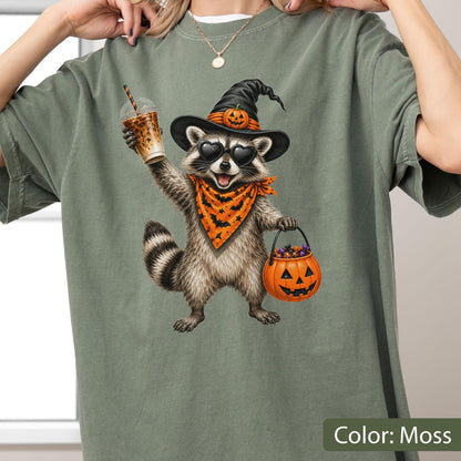 May include: A moss green t-shirt featuring a graphic of a raccoon wearing a black witch hat with a small pumpkin, heart-shaped sunglasses, and an orange bandana with black bats. The raccoon holds an iced coffee and a pumpkin-shaped candy bucket.