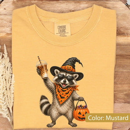 May include: A mustard yellow t-shirt featuring a graphic of a raccoon wearing a black witch hat with a small pumpkin, heart-shaped sunglasses, and an orange bat-patterned bandana. The raccoon holds an iced coffee and a jack-o'-lantern bucket.