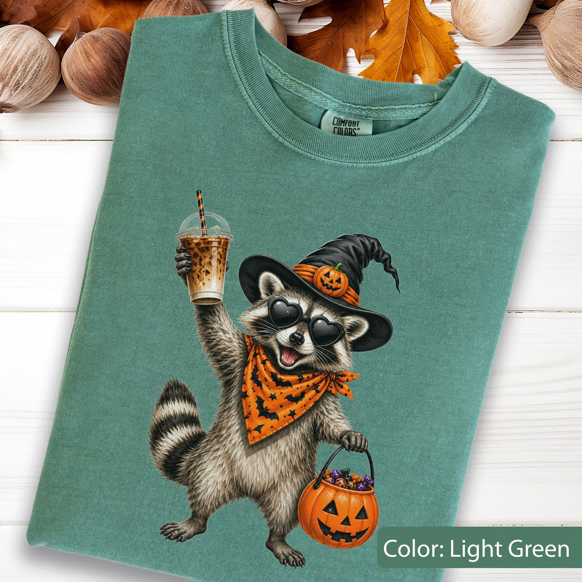 May include: A light green t-shirt featuring a graphic of a raccoon wearing a black witch hat with a small pumpkin, heart-shaped sunglasses, and an orange bandana with black bats. The raccoon holds an iced coffee and a jack-o'-lantern bucket.
