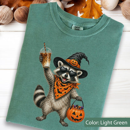 May include: A light green t-shirt featuring a graphic of a raccoon wearing a black witch hat with a small pumpkin, heart-shaped sunglasses, and an orange bandana with black bats. The raccoon holds an iced coffee and a jack-o'-lantern bucket.