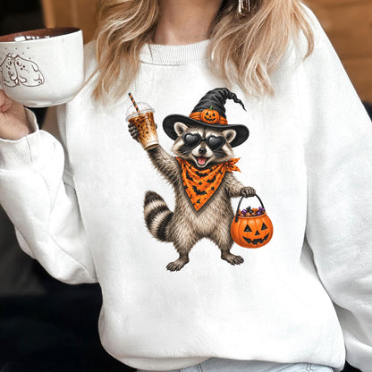 May include: A white sweatshirt featuring a graphic of a raccoon wearing a black witch hat with a small pumpkin, heart-shaped sunglasses, and an orange bandana with black bats. The raccoon holds an iced coffee and an orange jack-o'-lantern bucket.