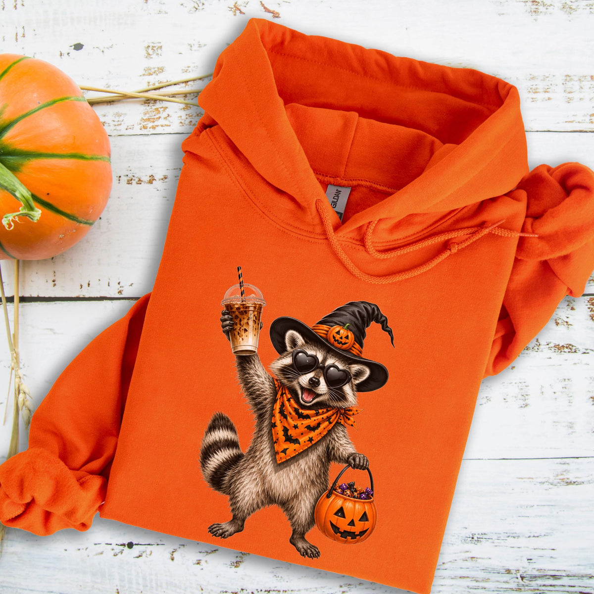 May include: An orange hooded sweatshirt featuring a graphic of a raccoon wearing a witch hat with a pumpkin, heart-shaped sunglasses, and an orange bandana with black bats. The raccoon holds an iced coffee and a jack-o'-lantern candy bucket.
