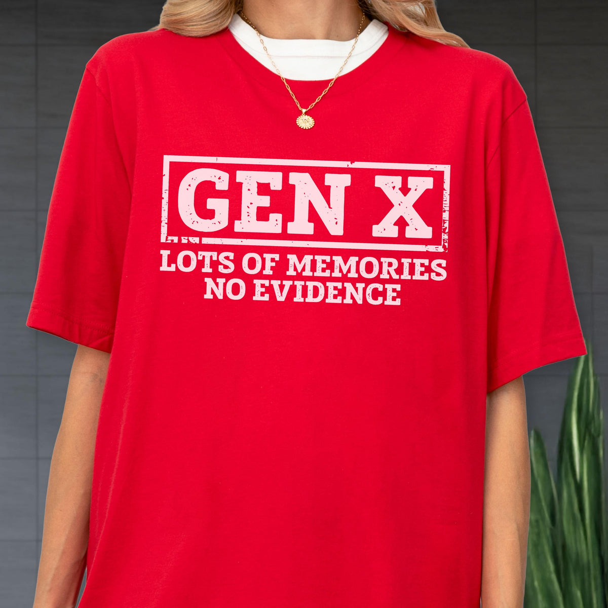 May include: A bright red short-sleeved t-shirt featuring a white rectangular graphic with the distressed text "GEN X" at the top and "LOTS OF MEMORIES NO EVIDENCE" underneath. The shirt is worn over a white crew neck base layer with a gold chain necklace.