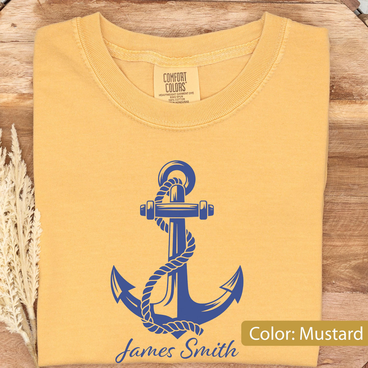 May include: A mustard yellow t-shirt featuring a blue graphic of a nautical anchor wrapped in rope. The shirt has a crew neckline and a Comfort Colors brand label at the inner collar. The text James Smith is printed in blue below the anchor.