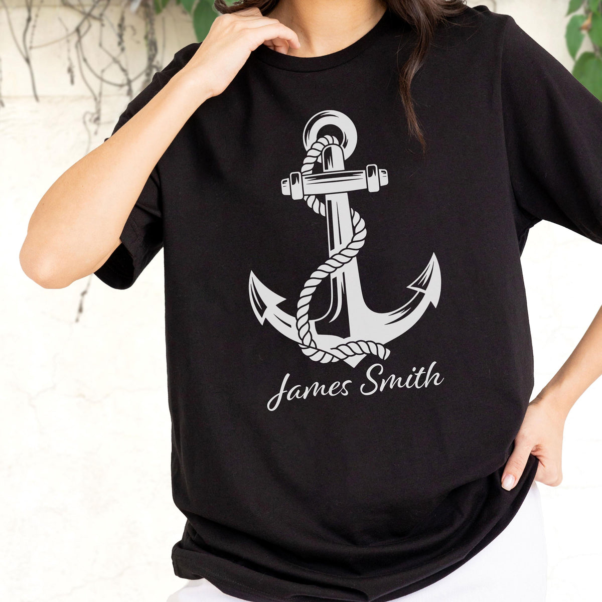 May include: A black short-sleeved t-shirt featuring a large, light gray graphic of a nautical anchor wrapped in a thick rope. Below the anchor, the name James Smith is printed in a white, elegant cursive script font across the chest area.