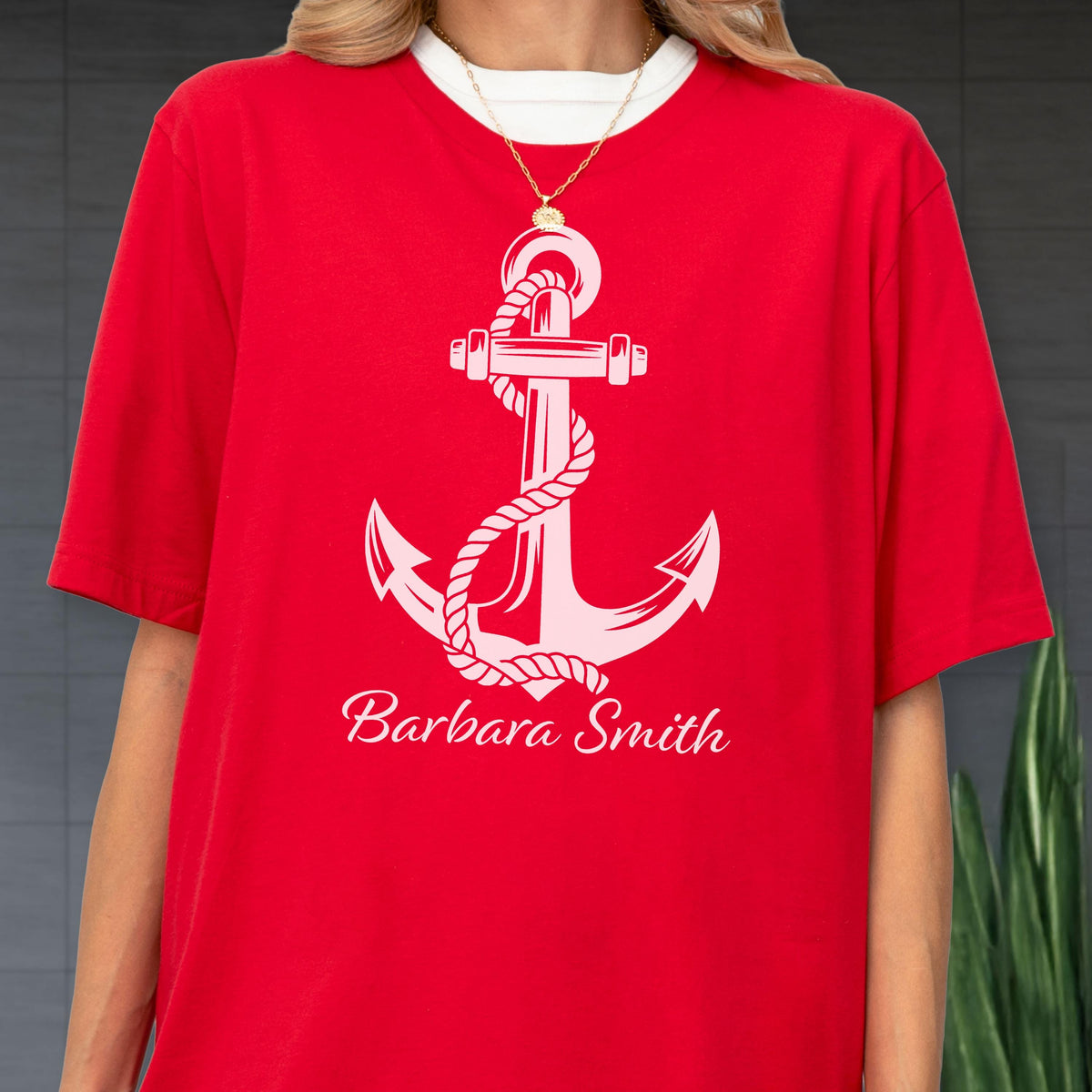 May include: A bright red short-sleeved t-shirt featuring a large white graphic of a nautical anchor wrapped in a rope. Below the anchor, the name Barbara Smith is printed in a white cursive script font. A thin gold chain necklace is visible.