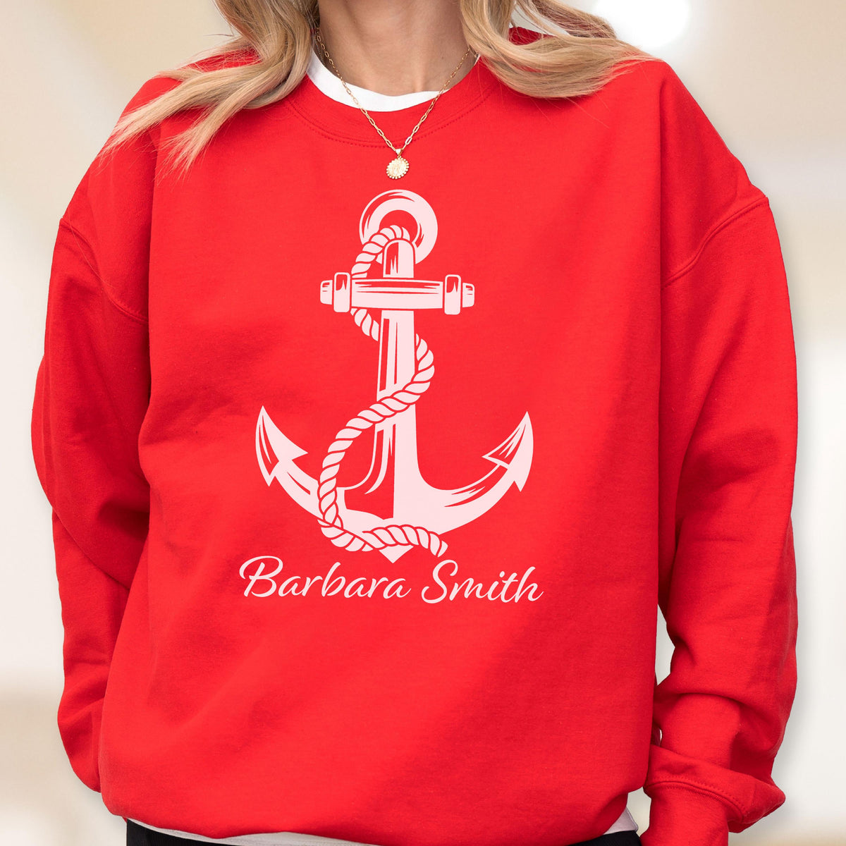 May include: A bright red crewneck sweatshirt featuring a large white graphic of a nautical anchor wrapped in rope. Below the anchor, the name Barbara Smith is printed in a white cursive script font. The garment has long sleeves and a ribbed neckline.