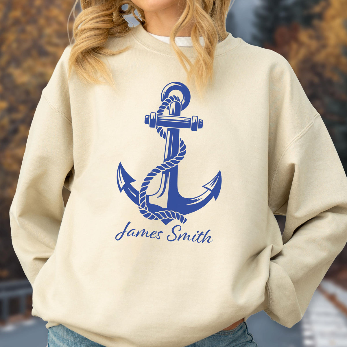 May include: A beige crewneck sweatshirt featuring a large blue graphic of a nautical anchor wrapped in a rope. Below the anchor, the name James Smith is printed in a blue script font. The garment has long sleeves and a ribbed neckline.