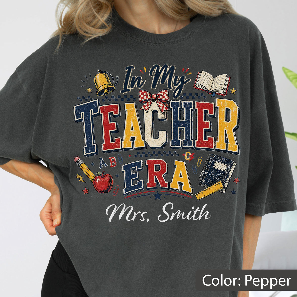 May include: A charcoal gray t-shirt featuring the text In My Teacher Era and Mrs. Smith. The graphic includes a gold bell, a red apple, a pencil, an open book, a spiral notebook, a ruler, and a red and white checkered bow in a distressed style.
