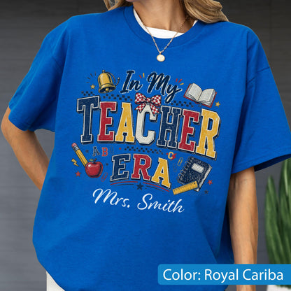 May include: A royal blue t-shirt featuring a graphic print with the text In My Teacher Era and Mrs. Smith. The design includes a gold bell, a red apple, a pencil, an open book, a spiral notebook, a ruler, and a red and white checkered bow.