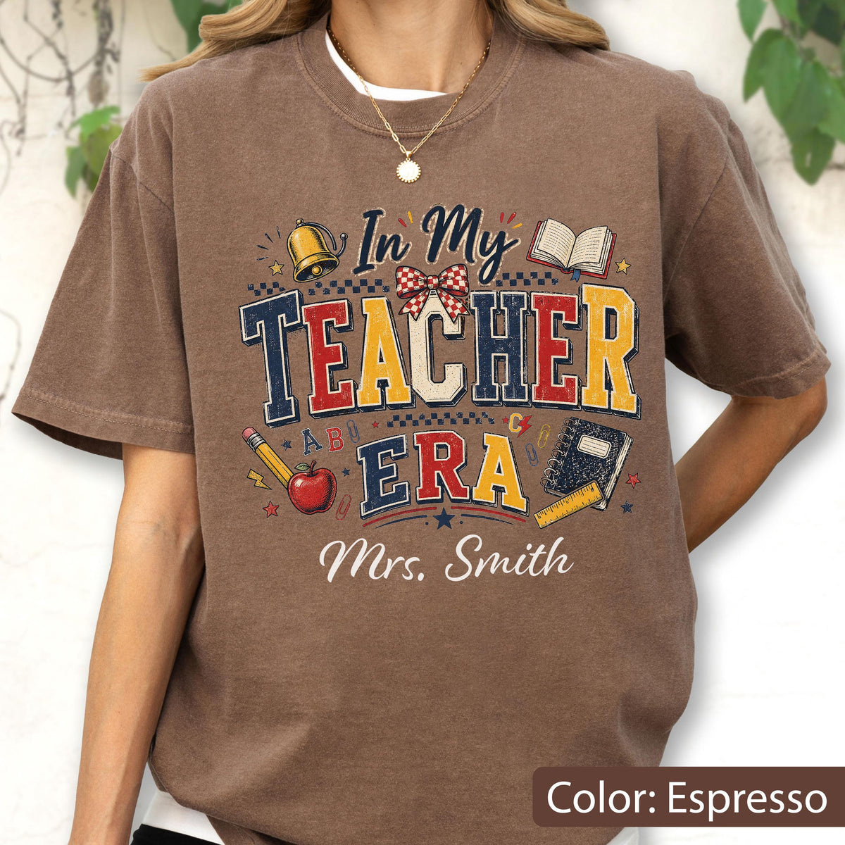 May include: An espresso-colored short-sleeve t-shirt featuring a graphic print with the text In My Teacher Era and Mrs. Smith. The design includes illustrations of a gold bell, a red apple, a pencil, a notebook, a ruler, and a red plaid bow.