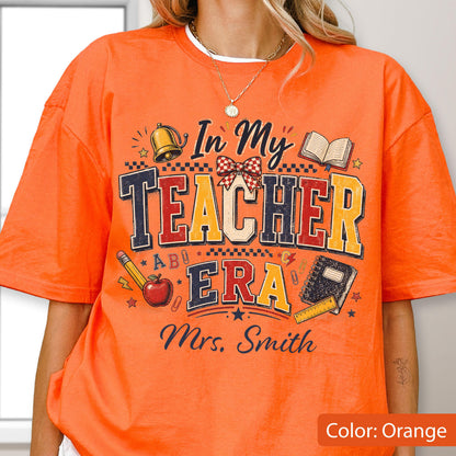 May include: An orange t-shirt featuring a graphic print with the text In My Teacher Era and Mrs. Smith. The design includes a gold bell, a red apple, a pencil, a notebook, a checkered bow, and small star icons in shades of red, yellow, and blue.