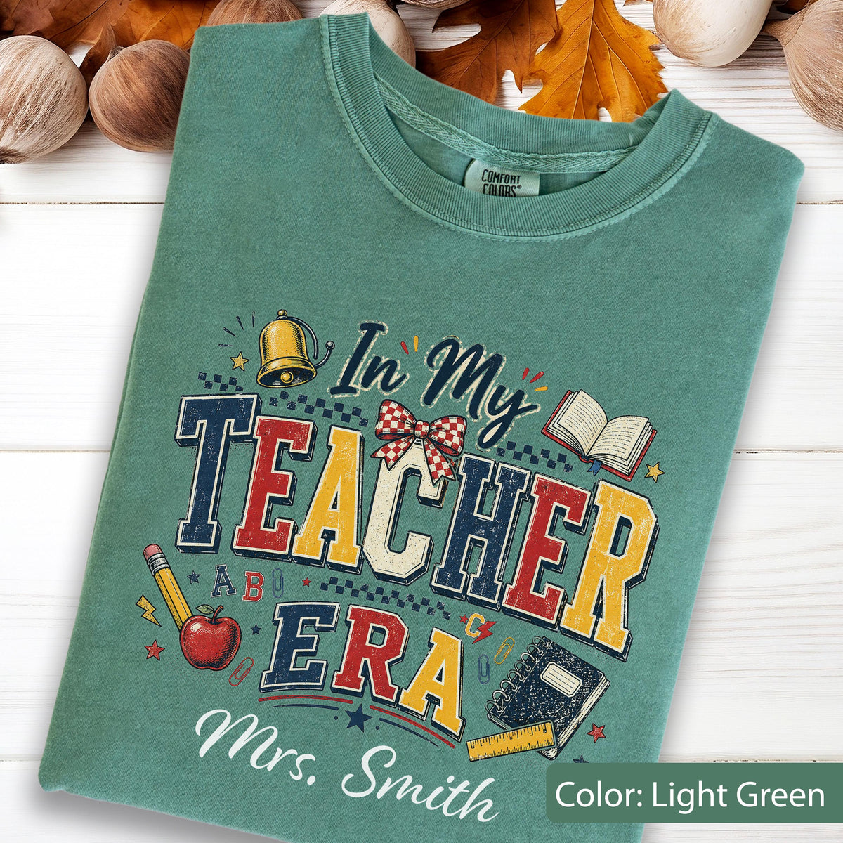 May include: A light green t-shirt featuring a graphic print with the text "In My TEACHER ERA" and "Mrs. Smith". The design includes illustrations of a gold bell, a red apple, a pencil, an open book, a notebook, a ruler, and a red plaid bow.