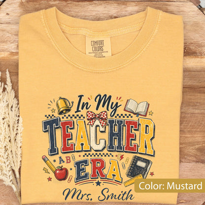 May include: A mustard yellow t-shirt featuring a graphic print with the text In My Teacher Era and Mrs. Smith. The design includes illustrations of a bell, an open book, a red apple, a notebook, a pencil, and a red and white checkered bow.