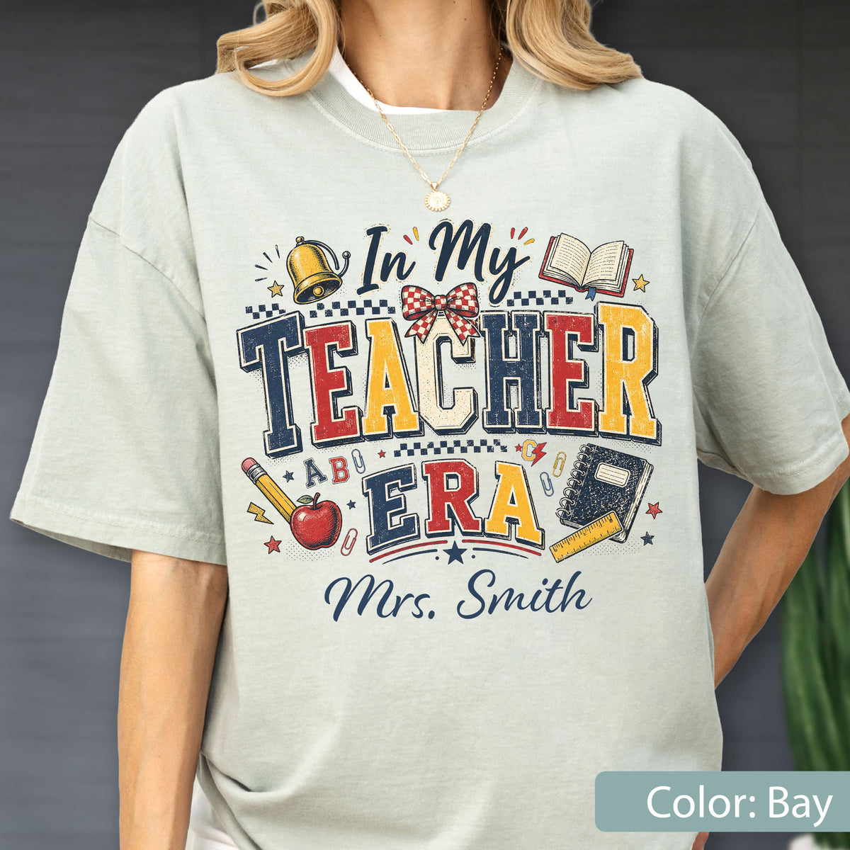 May include: A light sage green t-shirt featuring a graphic print with the text In My Teacher Era and Mrs. Smith. The design includes a gold bell, a red apple, a pencil, a notebook, a ruler, and a red and white checkered bow in a vintage style.