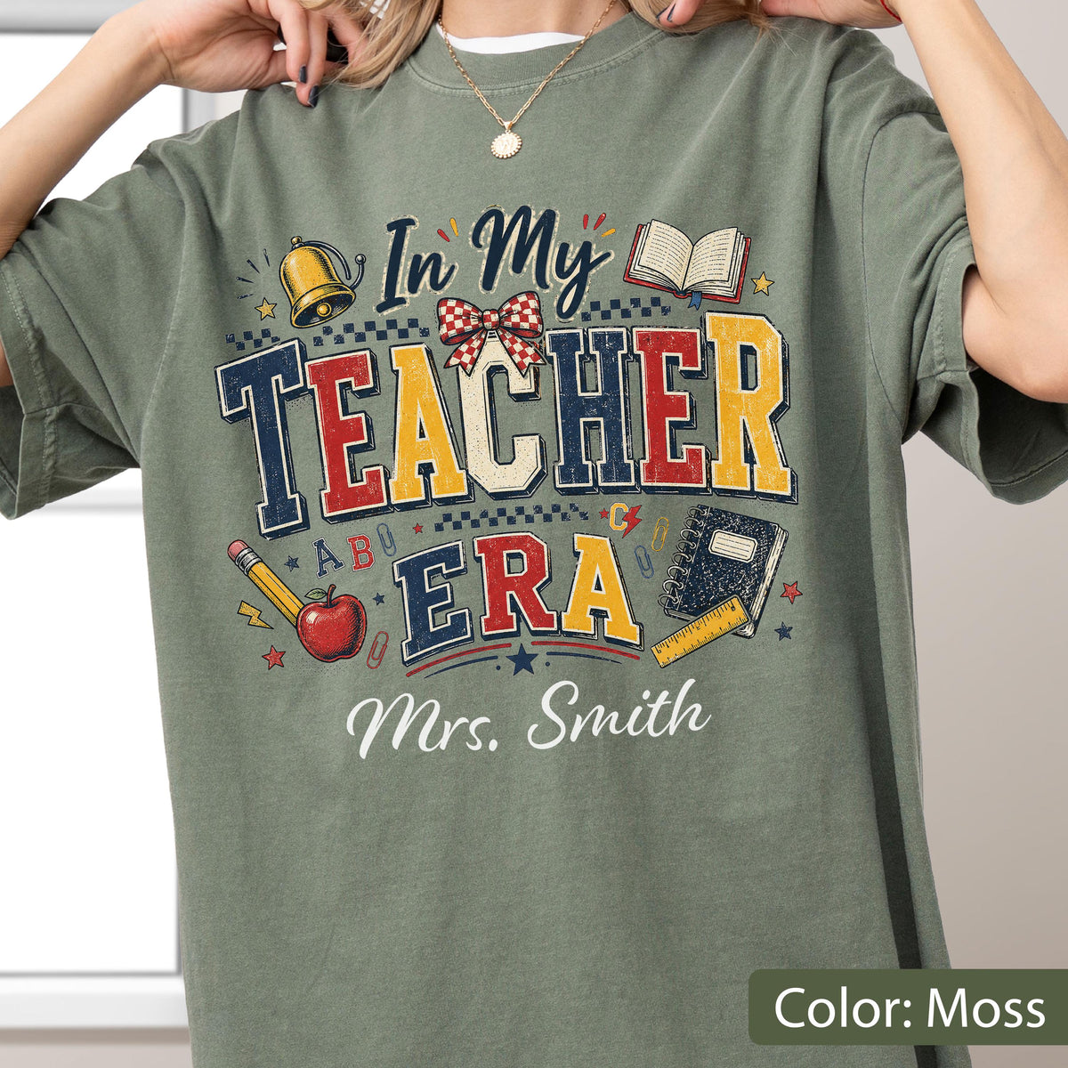 May include: A moss green t-shirt featuring a graphic print with the text In My Teacher Era and Mrs. Smith. The design includes a gold bell, an open book, a red apple, a pencil, a notebook, a ruler, and a red and white checkered bow.