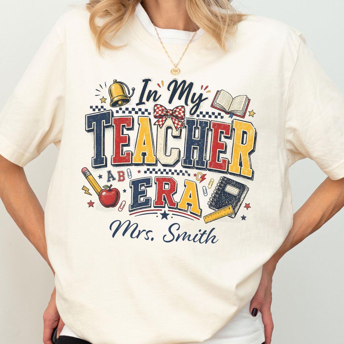 May include: A cream-colored t-shirt featuring a graphic design with the text In My Teacher Era and Mrs. Smith. The design includes illustrations of a gold bell, a red apple, a pencil, an open book, a notebook, a ruler, and a red plaid bow.