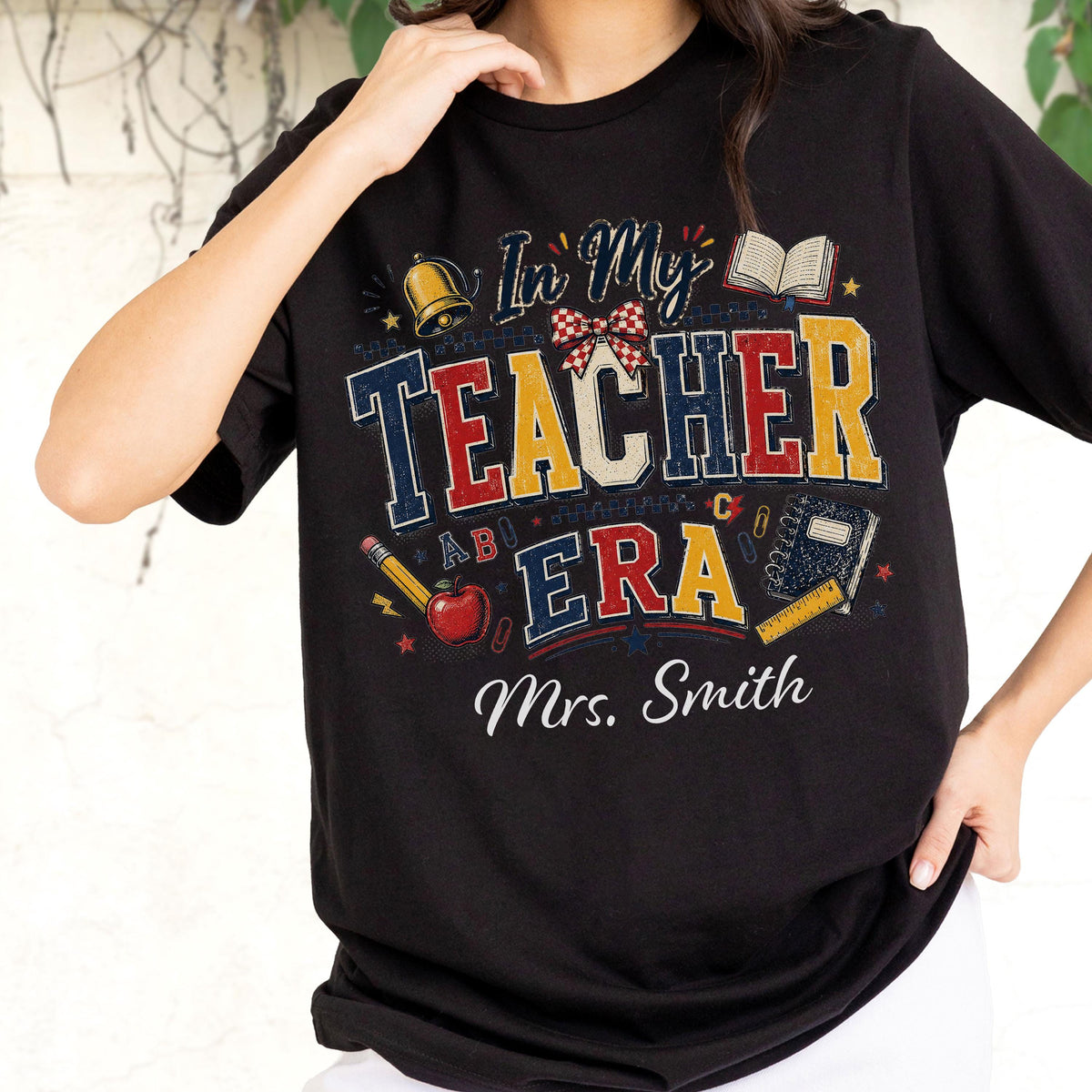 May include: A black short-sleeved t-shirt featuring a graphic print with the text In My Teacher Era and Mrs. Smith. The design includes a bell, an open book, a red apple, a pencil, a notebook, a ruler, and a red and white checkered bow.