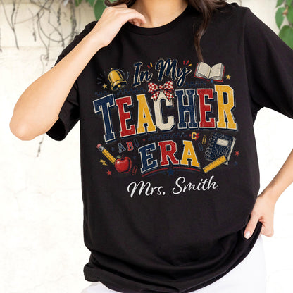 May include: A black short-sleeved t-shirt featuring a graphic print with the text In My Teacher Era and Mrs. Smith. The design includes a bell, an open book, a red apple, a pencil, a notebook, a ruler, and a red and white checkered bow.