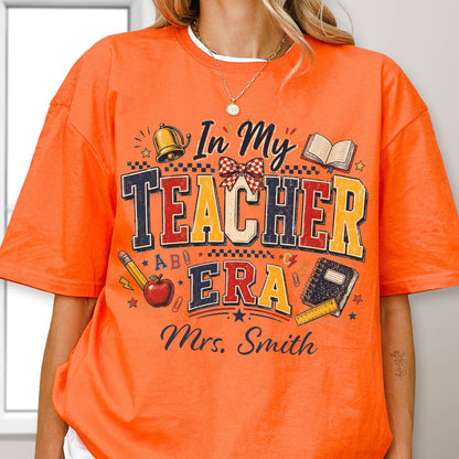 May include: An orange t-shirt featuring a graphic print with the text In My Teacher Era and Mrs. Smith. The design includes illustrations of a gold bell, a red apple, a pencil, a notebook, a ruler, a bow, and stars in blue, yellow, and red.
