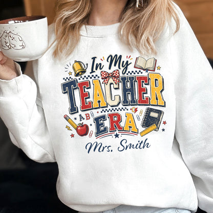 May include: A white crewneck sweatshirt featuring a graphic print with the text In My Teacher Era and Mrs. Smith. The design includes a gold bell, an open book, a red apple, a pencil, a notebook, a ruler, and a red and white checkered bow.