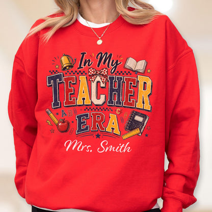 May include: A bright red crewneck sweatshirt featuring a graphic print with the text In My Teacher Era and Mrs. Smith. The design includes illustrations of a gold bell, an open book, a red apple, a pencil, a notebook, and a ruler with a bow.