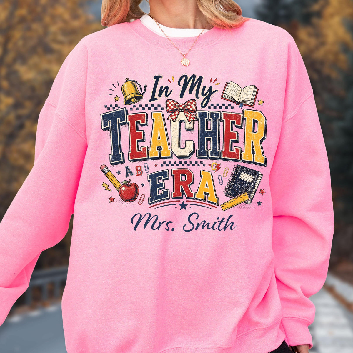 May include: A bright pink crewneck sweatshirt featuring a graphic print with the text In My Teacher Era and Mrs. Smith. The design includes illustrations of a bell, an open book, a red apple, a pencil, a notebook, a ruler, and a checkered bow.