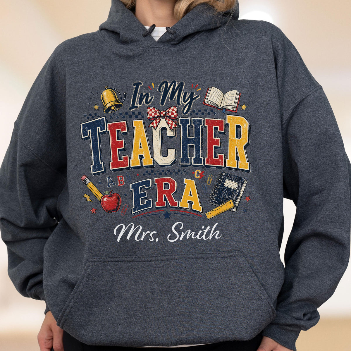May include: A dark heather gray hooded sweatshirt featuring a graphic print with the text In My TEACHER ERA and Mrs. Smith. The design includes a gold bell, an open book, a red apple, a yellow pencil, a blue notebook, and a red plaid bow.