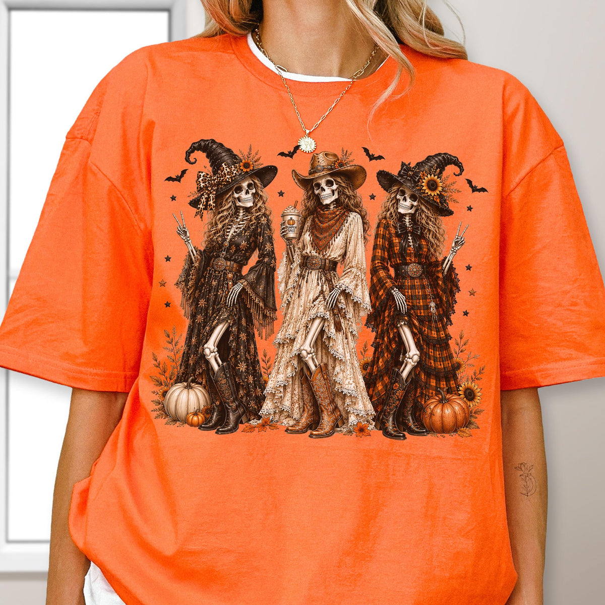May include: A bright orange t-shirt featuring a graphic of three skeletons dressed in western-style clothing, including hats, long dresses, and boots. The skeletons are surrounded by pumpkins, sunflowers, and bats, with a coffee cup held by the center figure.