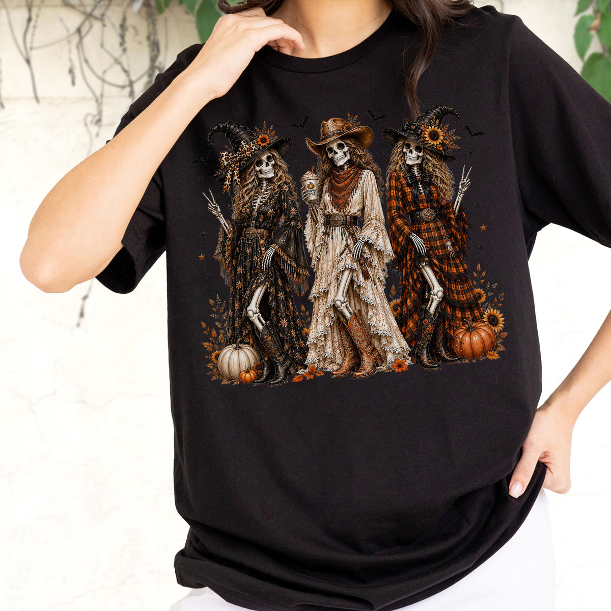 May include: A black short-sleeve t-shirt featuring a graphic print of three skeletons dressed in western-style clothing, including hats, long dresses, and boots. The skeletons are surrounded by pumpkins, sunflowers, and small bats on a dark background.