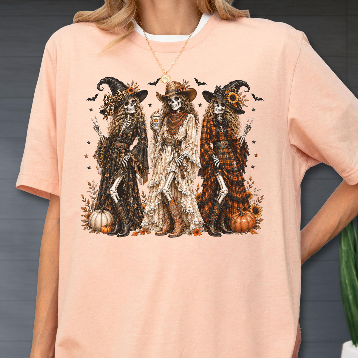 May include: A peach-colored t-shirt featuring a graphic print of three skeletons dressed in western-style clothing, including hats, long dresses, and boots. The skeletons are surrounded by pumpkins, sunflowers, and small bats on a light background.
