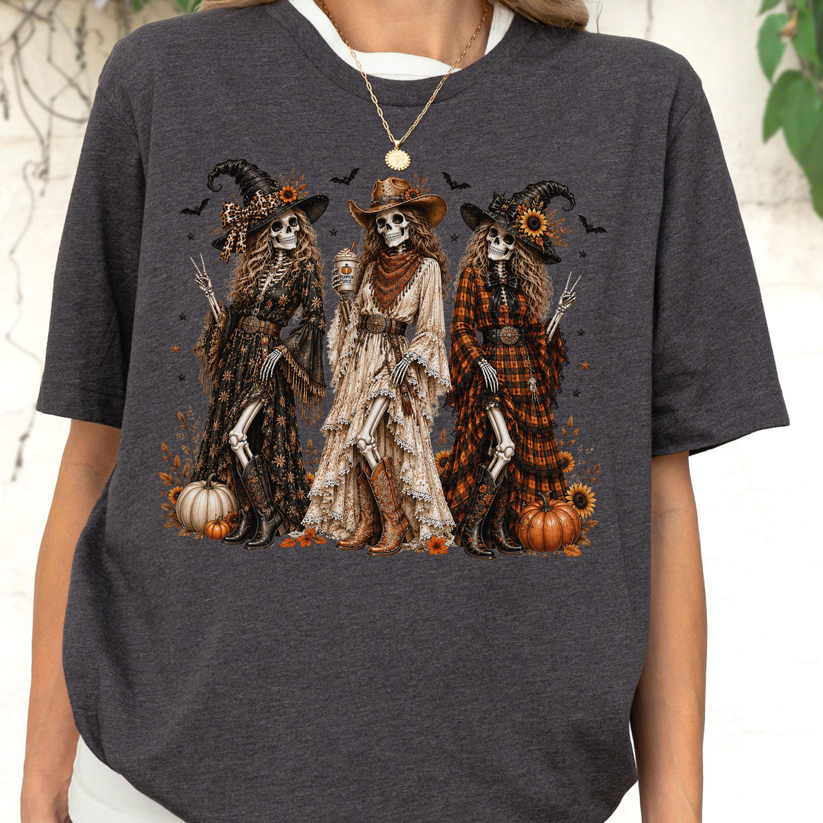 May include: A dark charcoal gray t-shirt featuring a graphic print of three skeletons dressed in western-style clothing, including hats, long dresses, and boots. The skeletons are surrounded by sunflowers, pumpkins, and small bats on a dark background.