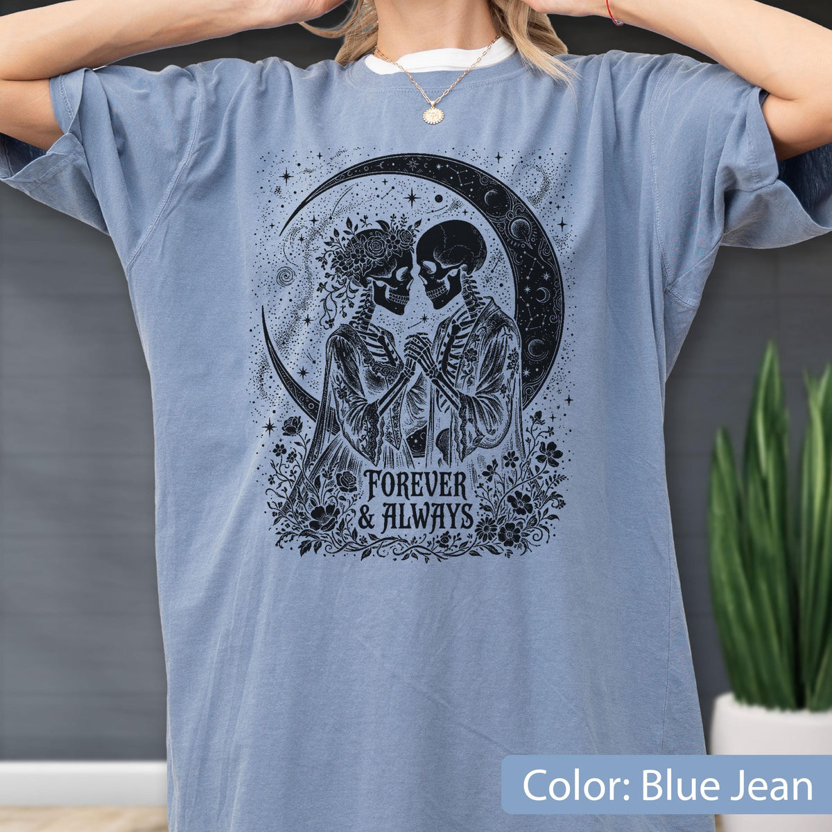 May include: A blue jean colored t-shirt featuring a black graphic print of two skeletons holding hands under a crescent moon. The design includes floral elements and the text "FOREVER & ALWAYS" centered below the skeletons in a decorative font.