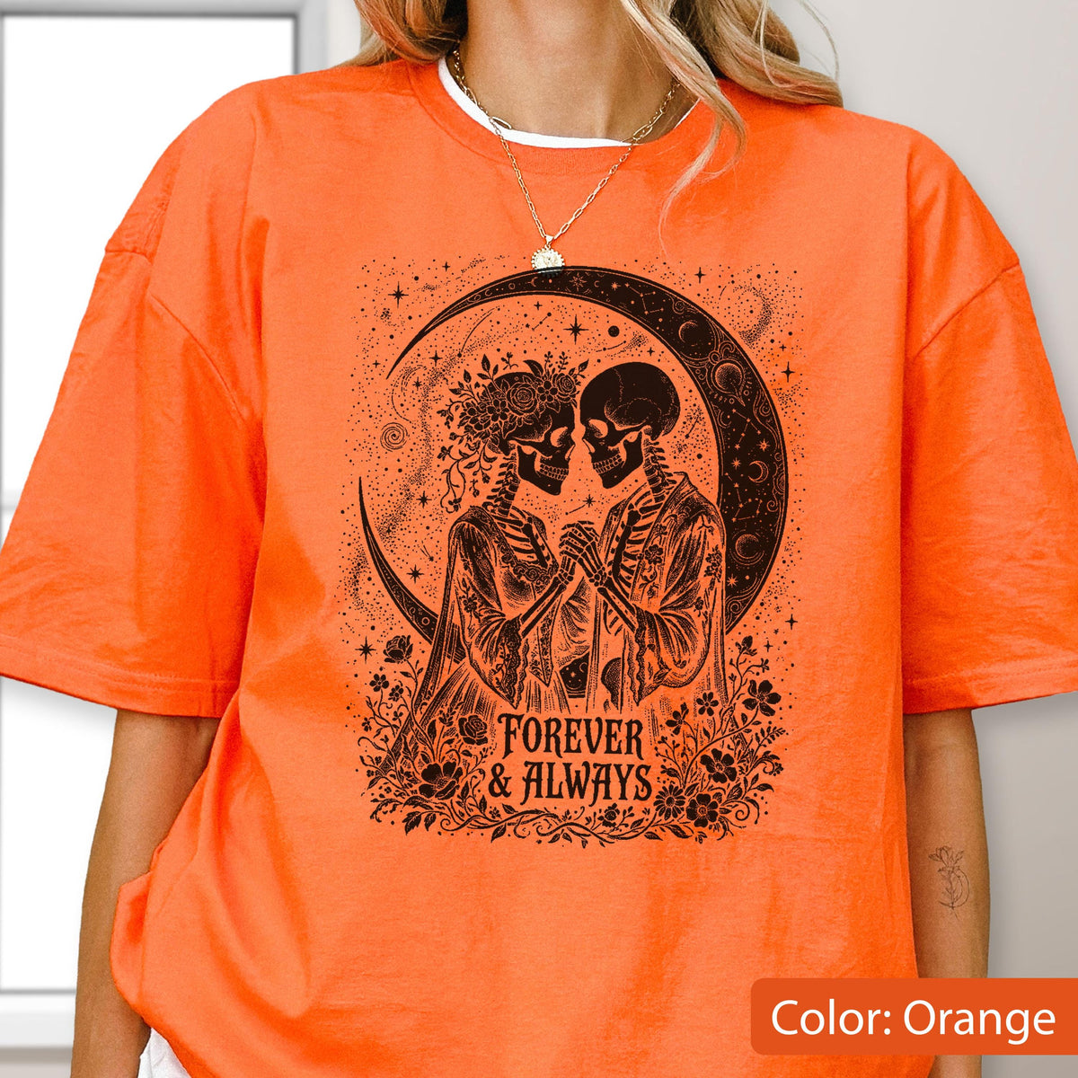 May include: An orange t-shirt featuring a black graphic print of two skeletons holding hands under a crescent moon. The skeletons are surrounded by floral elements and stars. The text "FOREVER & ALWAYS" is printed in a decorative font below them.